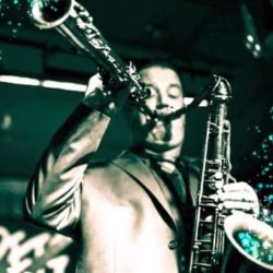 A man playing the saxophone in front of trees.